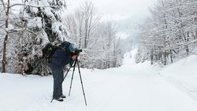 A male photographer adjusts his camera on a tripod to shoot the edge of a mountain forest in the beautiful Carpathians in snowy weather