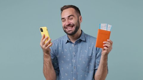 Joyful excited fun young brunet bearded man 20s wears denim shirt hold use mobile cell phone doing online shopping hold passport boarding tickets booking tour isolated on pastel light blue background