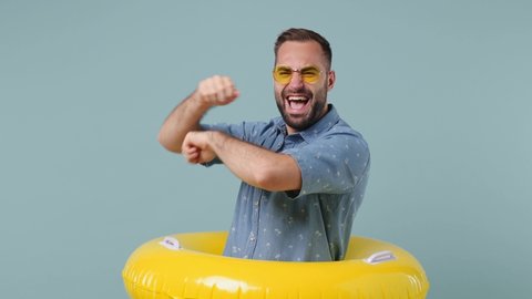 Traveler tourist fun young brunet bearded man 20s wears denim shirt yellow sunglasses stand in inflatable ring dance isolated on pastel light blue background. Summer vacation sea rest sun tan concept