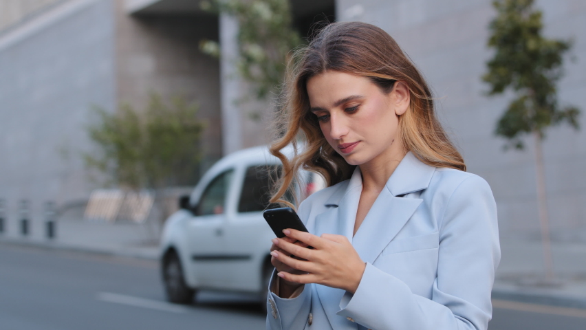 Serious caucasian young elegant business woman using social media application on smartphone texting messages receive news typing with mobile phone outdoors online chatting technology browsing in city