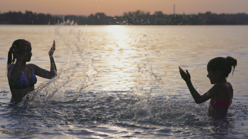 Two funny funny sisters splash on each other with shiny transparent clear water in a blue shallow lake against the backdrop of a bright sunny golden summer warm sunset. UHD video slow-motion 4K