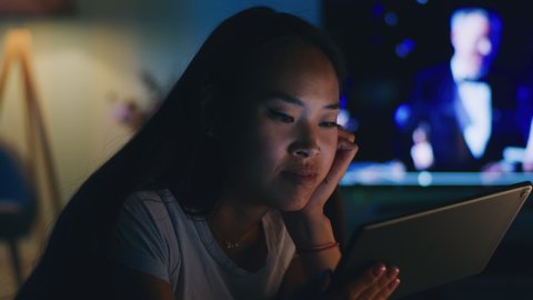 Pan around view of Asian woman leaning on hand and watching video on tablet while resting near TV in dark living room at home