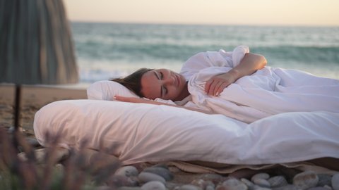 Happy young smiling woman sleeping in white soft bed with turquoise sea waves splashing with foam on sandy beach at background. Portrait of carefree Caucasian lady with closed eyes lying outdoors