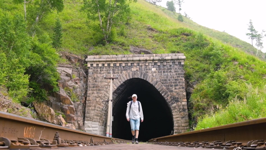 A male tourist with backpack walks along the rails of railway laid through tunnel. Slow motion. Local travel, exploring new places and hiking on weekends. Active recreation, the concept of tourism.