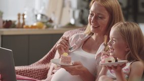 Happy pregnant woman and cute little daughter sitting together on sofa at home, eating desserts, watching movie online and laughing