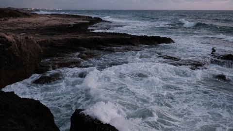 A rocky, wild beach, where waves hits the rocks vigorously at twilight while distant city lights are visible at the horizon. a 4K video clip, Achziv Beach, Israel.