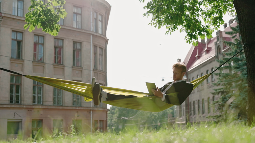 Caucasian redhead businessman lying in yellow hammock at city park and surfing internet on digital tablet. Successful male checking working emails outdoors.