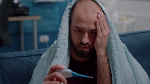 Close up of unwell adult looking at thermometer and measuring temperature while wrapped in blanket. Sick man with illness holding medical instrument to check fever and symptoms.