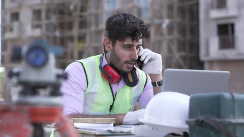 Indian civil engineer talking on smartphone while working on laptop at construction site.Worker wearing earmuff at construction site outdoor.Asian male wearing safety jacket and talking on mobile.
