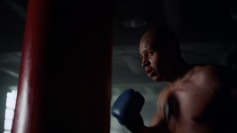 Male professional boxer practicing kicks in dark gym. African american man boxing  punch bag. Portrait of active sport man training punches. Shirtless sportsman box exercise in sport club. Strength