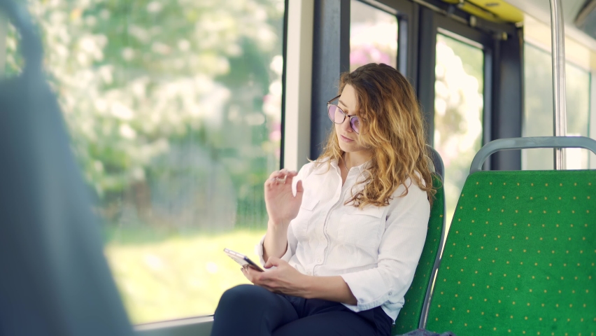 Caucasian young woman rides public transport, sits on a seat use a mobile phone, smartphone by the bus window. Portrait Business lady in glasses and a jacket or female student traveling inside bus | Shutterstock HD Video #1079246561