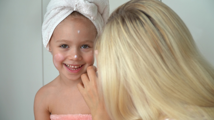 Mother applies cream to daughters face after bathing in bathroom. Little child with wrapped towel on head looks at camera and laughs. Baby skin care Royalty-Free Stock Footage #1079247005