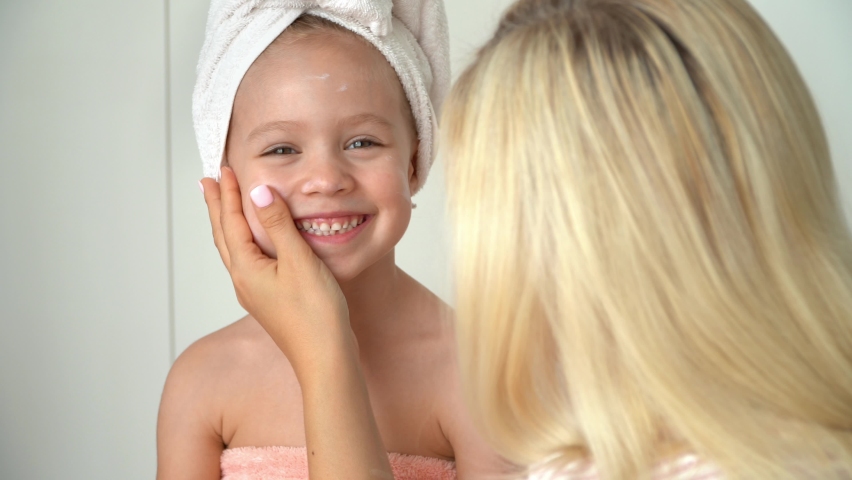 Mother applies cream to daughters face after bathing in bathroom. Little child with wrapped towel on head looks at camera and laughs. Baby skin care | Shutterstock HD Video #1079247005