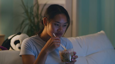 Asian female eating noodles from plastic jar and watching TV while chilling on couch in evening at home