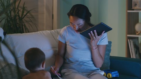 Asian woman eating noodles and talking on tablet while sitting on couch near son in obscure living room in evening at home