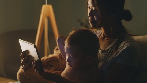 Asian woman and cute boy resting on sofa and watching interesting cartoon on tablet together in obscure living room at night at home