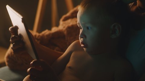 Cute Asian boy watching video on tablet while resting in arms of crop mother at night at home