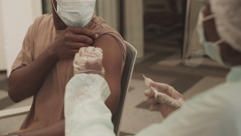 Over-the-shoulder medium shot of African American man getting Covid-19 vaccine shot in his arm sitting at modern doctor office