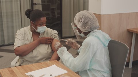 Medium shot of female medical worker in protective clothing, face shield and mask doing Covid-19 vaccine injection to African American woman at modern doctor office