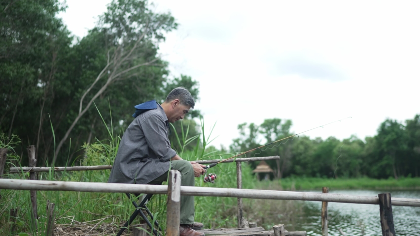 Elderly Asian man, gray hair, hat, holding a Catch rod, sits and sleeps fishing. On the edge of the lake, on vacation, retirement, hobbies for the elderly, concept