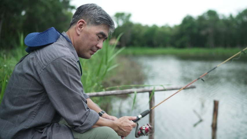 Elderly Asian man, gray hair, hat, holding a Catch rod, sits and sleeps fishing. On the edge of the lake, on vacation, retirement, hobbies for the elderly, concept