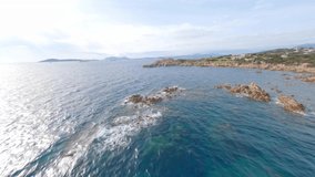 FPV video, view from above, stunning aerial view from an FPV drone flying at high speed over a green and rocky coastline bathed by a blue water. Costa Smeralda, Sardinia, Italy.