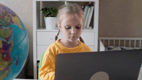 Adorable small child girl sitting at table, web surfing educational information online on computer, enjoying distant e-learning process, studying remotely from home, quarantine lifestyle concept