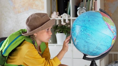 Child in a hat with a backpack dreams of a trip. Schoolgirl girl examines the globe through a magnifying glass. Tourism and travel concept. Closed borders