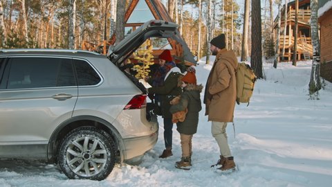 Handheld tracking of family of three wearing warm winter clothes taking backpack out of trunk of car and walking towards cozy A-frame cabin in forest