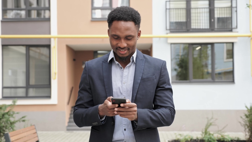 Black Man dressed in Business suit walking down the street and Reading something on his Phone. Green trees and Colorful Building on the background of Business Man which is raising his head. Royalty-Free Stock Footage #1079282405