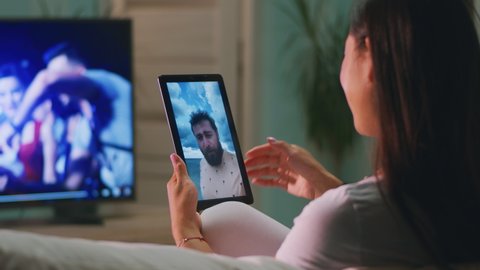 Cheerful woman smiling and speaking with bearded husband via digital tablet while resting on sofa near TV and making video call at home