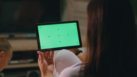Unrecognizable woman watching video on modern tablet with chromakey green screen while resting in living room near playing son