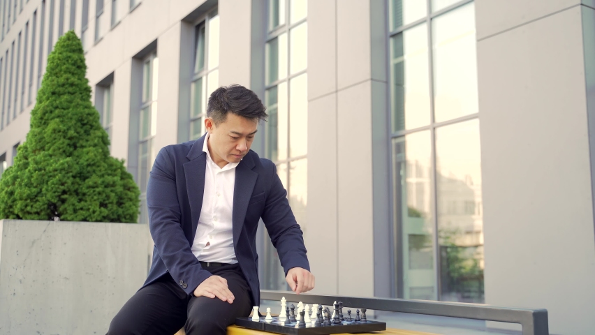Concentrated Young Asian businessman playing chess on a bench on an urban background of a modern office building. Business man making a move with chess. concept strategy competition and team work