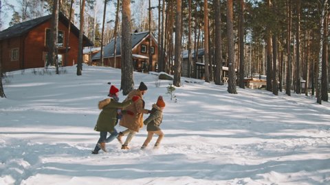 Slowmo PAN of happy parents and little girl in warm parkas, hats and scarfs running in snow on nice winter day. Cozy cottages surrounded by trees in background Arkivvideo