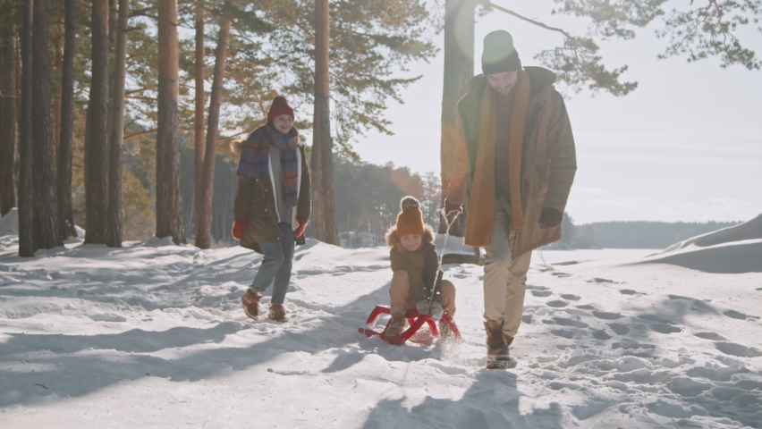 Slowmo tracking shot of happy man pulling sled with laughing little girl and running in snow while cheerful woman following them and smiling. Family of three enjoying winter day in forest Royalty-Free Stock Footage #1079284322