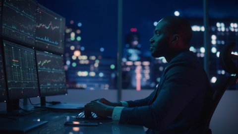 Financial Analyst Working on Computer with Multi-Monitor Workstation with Real-Time Stocks, Commodities and Exchange Market Charts. African American Businessman Works in Investment Bank Late at Night.