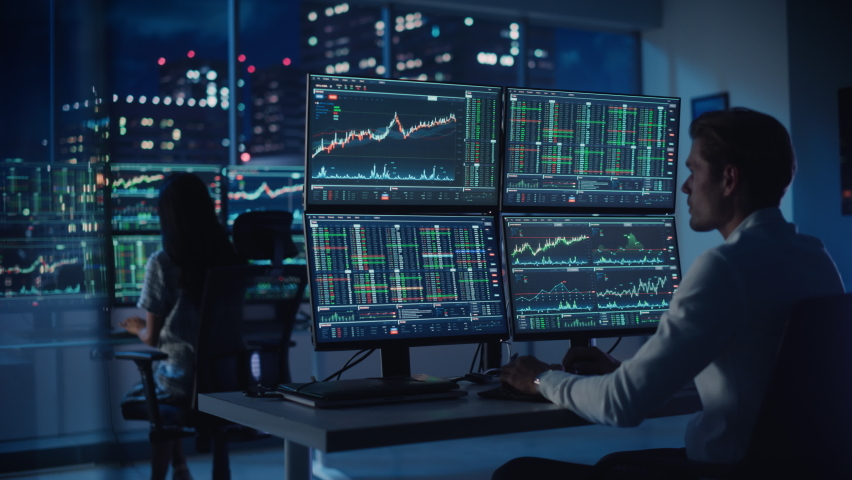 Financial Analyst Working on a Computer with Multi-Monitor Workstation with Real-Time Stocks, Commodities and Foreign Exchange Charts. Businessman Works in Investment Bank City Office at Night.