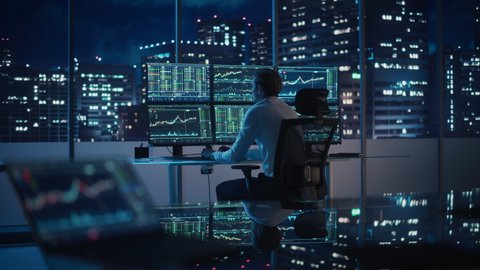 Financial Analyst Working on a Computer with Multi-Monitor Workstation with Real-Time Stocks, Commodities and Exchange Market Charts. Businessman Works in Investment Bank Downtown Office at Night.