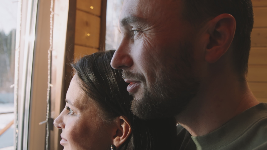 Handheld close up shot with slowmo of happy man and woman standing together by window and enjoying sunny winter day in their cottage