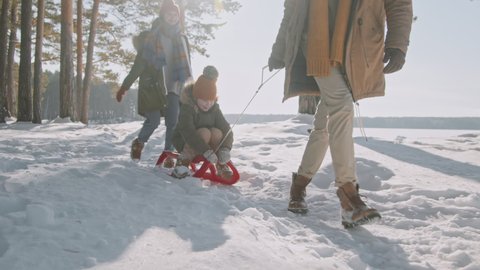 Slowmo tracking of father in warm winter clothes pulling cute 8-year-old girl on sled in forest. Cheerful mother walking behind them