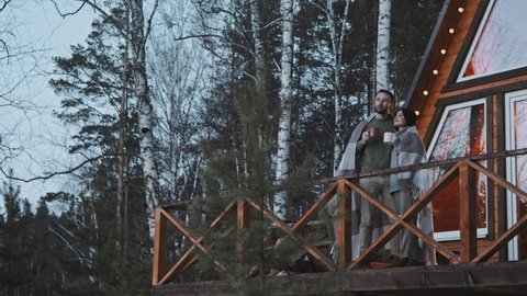 PAN slowmo shot of young man and woman standing wrapped in warm blanket on front porch of A-frame cabin and enjoying hot tea on winter evening