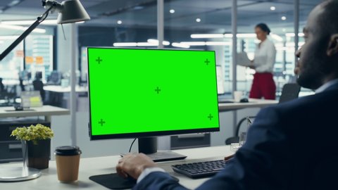 Successful Black Businessman Sitting at Desk Working on Green Screen Laptop Computer in Office. African American Businessperson using Chroma Key Display. Over Shoulder Static Shot