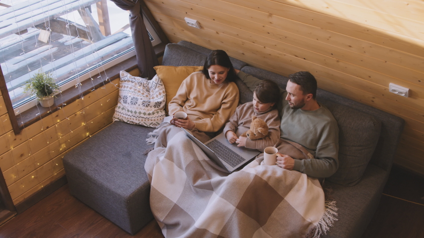 High angle slowmo of father, mother and daughter sitting together on couch with their legs wrapped in blanket. They are watching movie on laptop and drinking hot tea in their cozy cabin on winter day Royalty-Free Stock Footage #1079295557