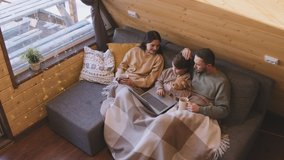 High angle slowmo of father, mother and daughter sitting together on couch with their legs wrapped in blanket. They are watching movie on laptop and drinking hot tea in their cozy cabin on winter day - Powered by Shutterstock - Get 15% off with code: PIKWIZARD15