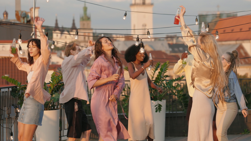Cheerful multicultural women and men dancing with hands above head on rooftop party. Happy friends drinking cocktails and enjoying favorite music. Concept of youth lifestyles. Royalty-Free Stock Footage #1079300276
