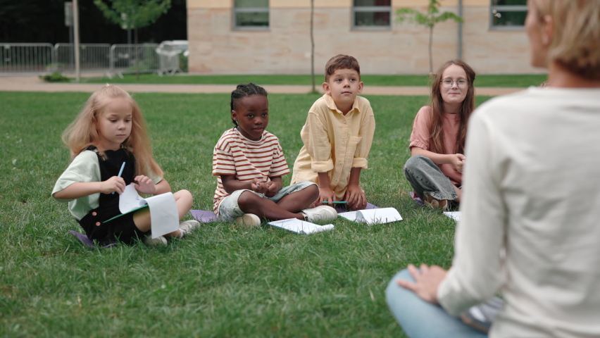 Group of diverse school children sitting on grass and raising their hands during lesson outdoors. Back view of female tutor giving various questions to curious kids on schoolyard. Royalty-Free Stock Footage #1079300306