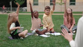 Group of diverse school children sitting on grass and raising their hands during lesson outdoors. Back view of female tutor giving various questions to curious kids on schoolyard. - Powered by Shutterstock - Get 15% off with code: PIKWIZARD15