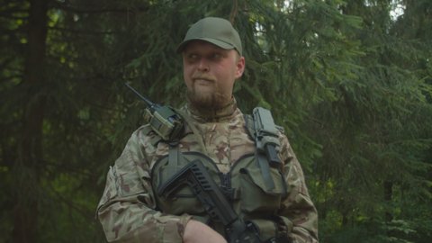 Portrait of adult bearded soldier in combat gear with firearm looking around outdoors. Fearless military man looking at camera, smirking with self-satisfaction in forest area