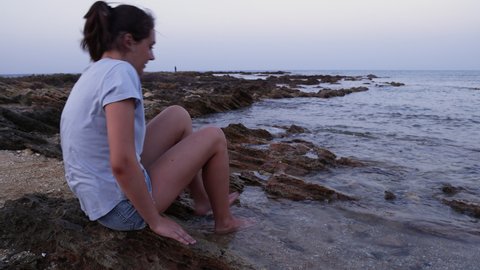 Teenage girl sitting on cliff by the sea at sunset, has legs in water, touch water by hand and looking down, wearing blue t-shirt and denim shorts, side view