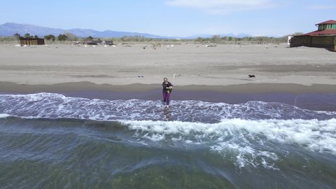 Young blonde woman in jeans and flannel shirt goes into sea, carrying dachshund dog in her arms. She lowers pet into water so that it learns to swim during training, shooting with drone from above